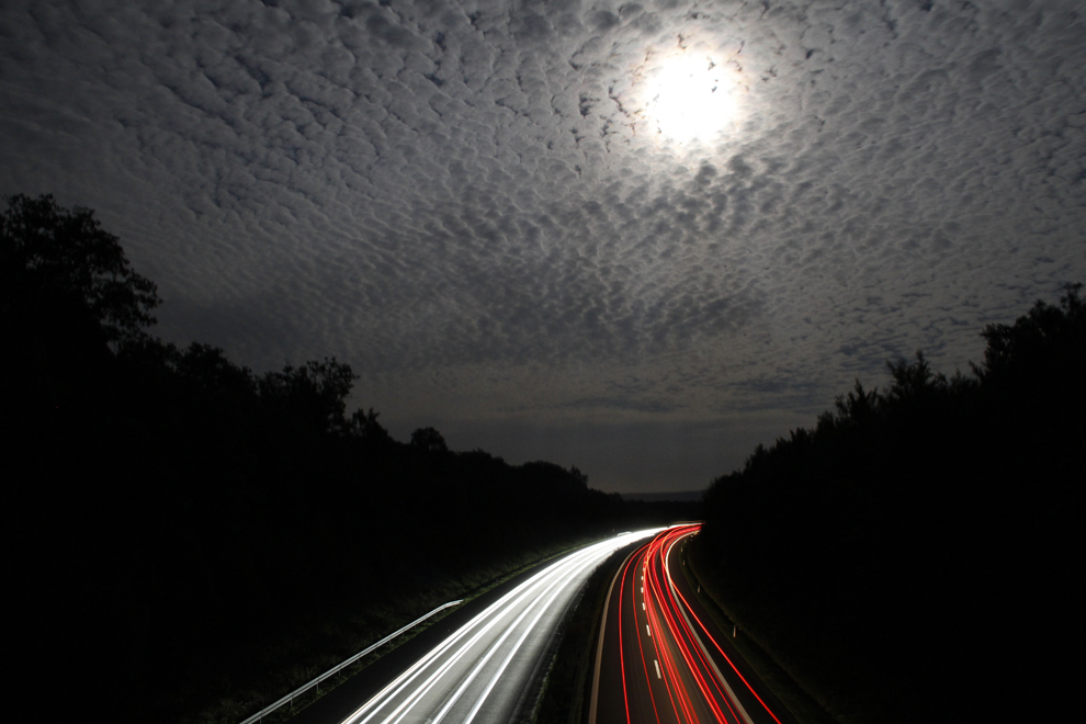 Fotografie cu expunere lungă: Luna plină poate fi vazută prin cerul înnorat în timp ce maşinile merg pe autostrada A70, în apropiere de Heinert, Germania.