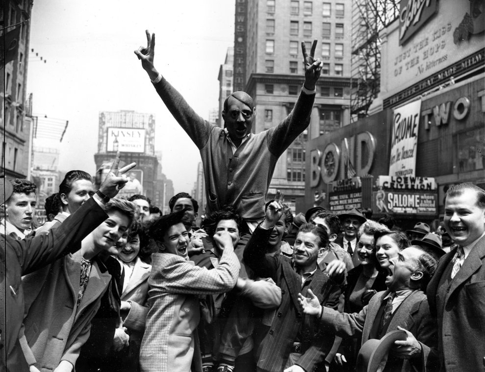 Americanii sărbătoresc capitularea Germaniei, în New York, strânşi în jurul unui tânăr cu masca lui Adolf Hitler, 7 mai 1945. (AP Photo/Harry Harris)