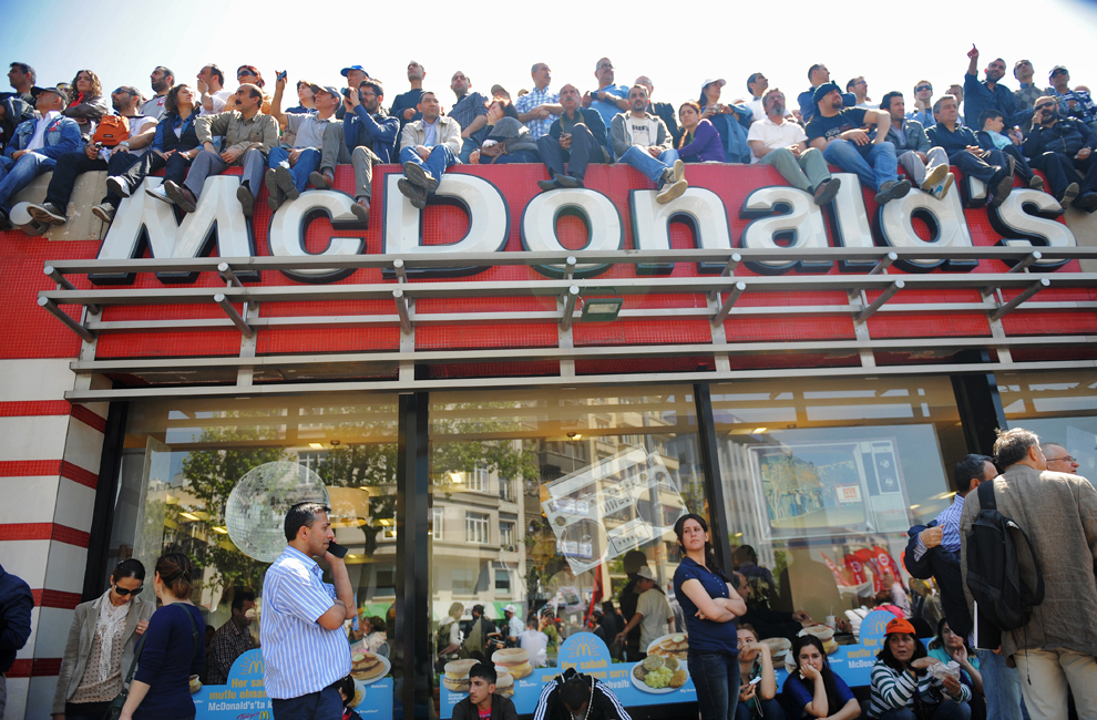 Persoane ce stau aşezate pe acoperişul unui restaurant McDonald’s urmăresc un miting organizat cu ocazia Zilei Internaţionale a Muncitorilor, în Istambul, Turcia, marţi, 1 mai 2012. 