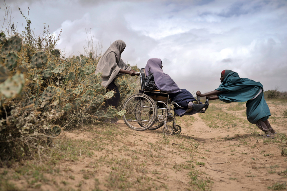 Doua femei o ajută pe o a treia, aflată într-un scaun cu rotile, sa treacă printr-un gard viu din cactuşi, la un centru de distribuţie a ajutoarelor, în Afgoye, Somalia, duminică, 4 august 2013. 